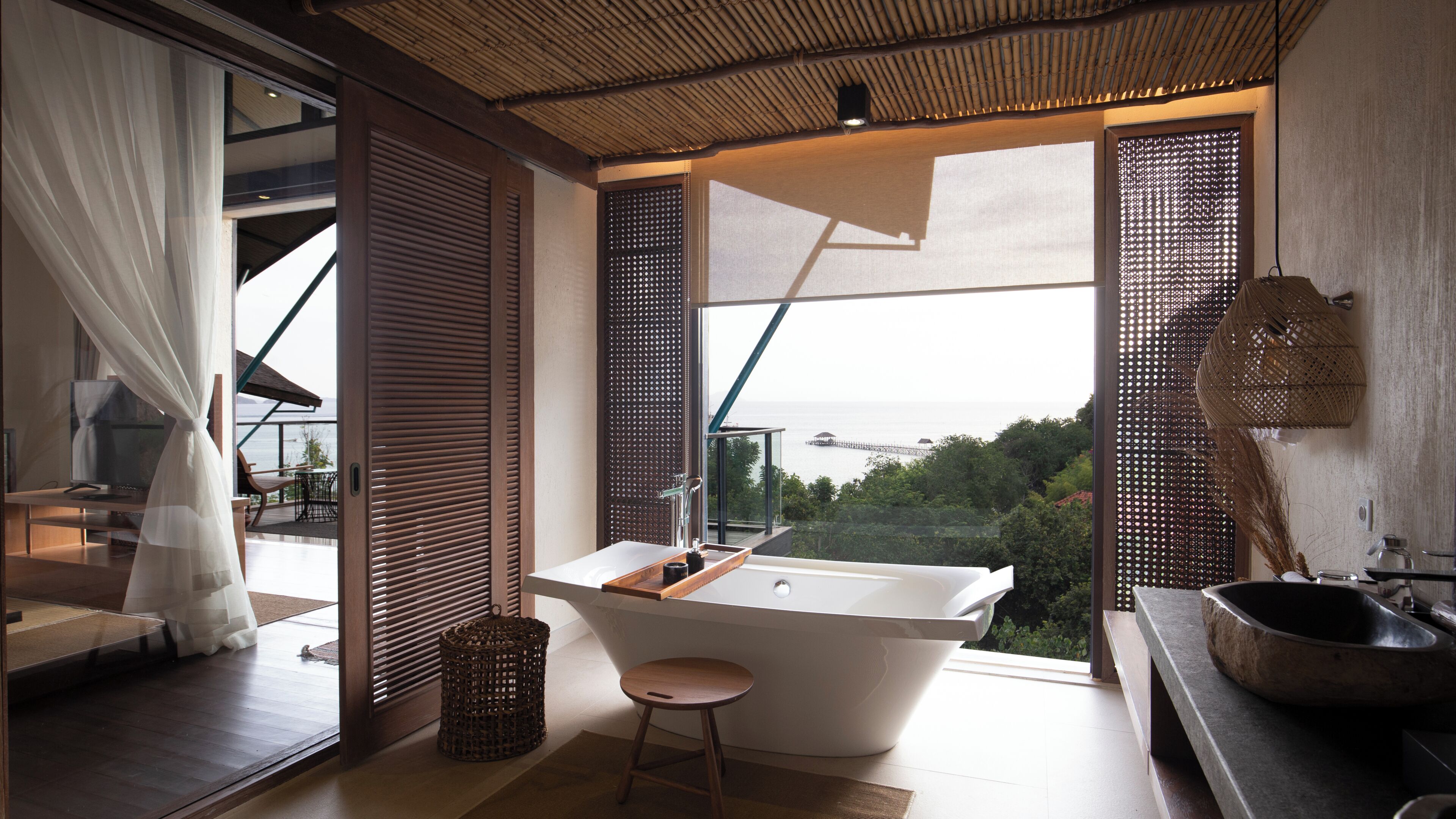 A modern bathroom with a freestanding bathtub, a wooden stool, and a view of the sea.