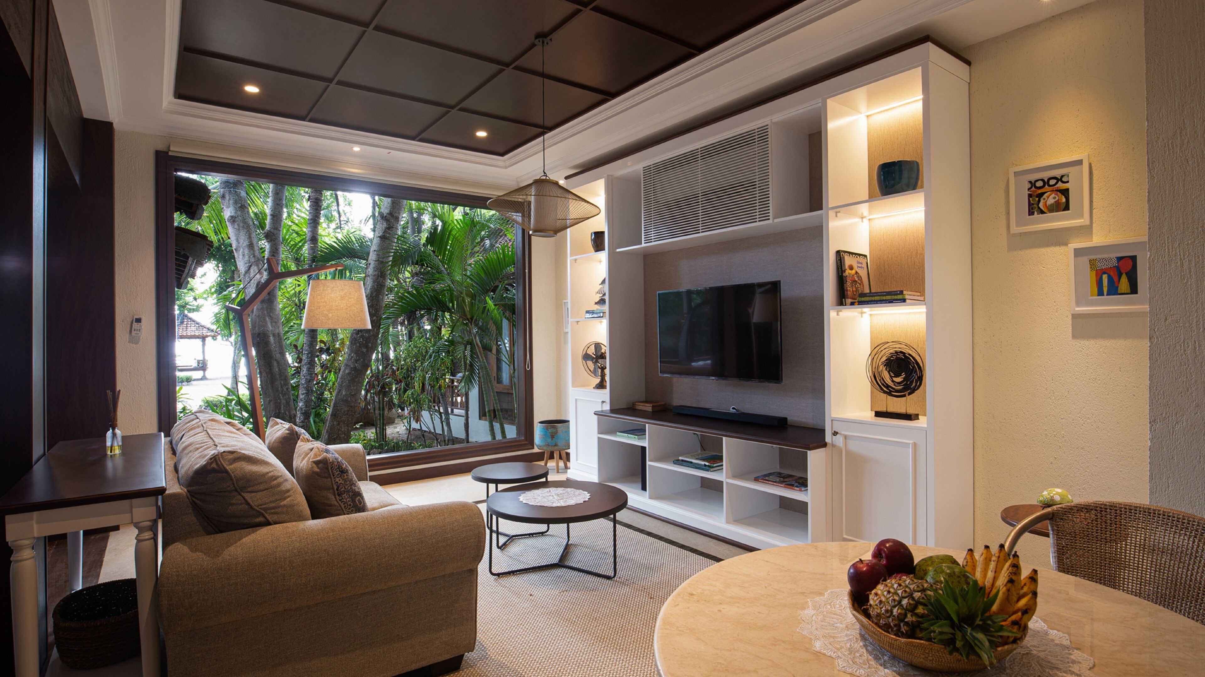 A modern living room with a sofa, coffee table, and TV. There is a large window with a view of trees.