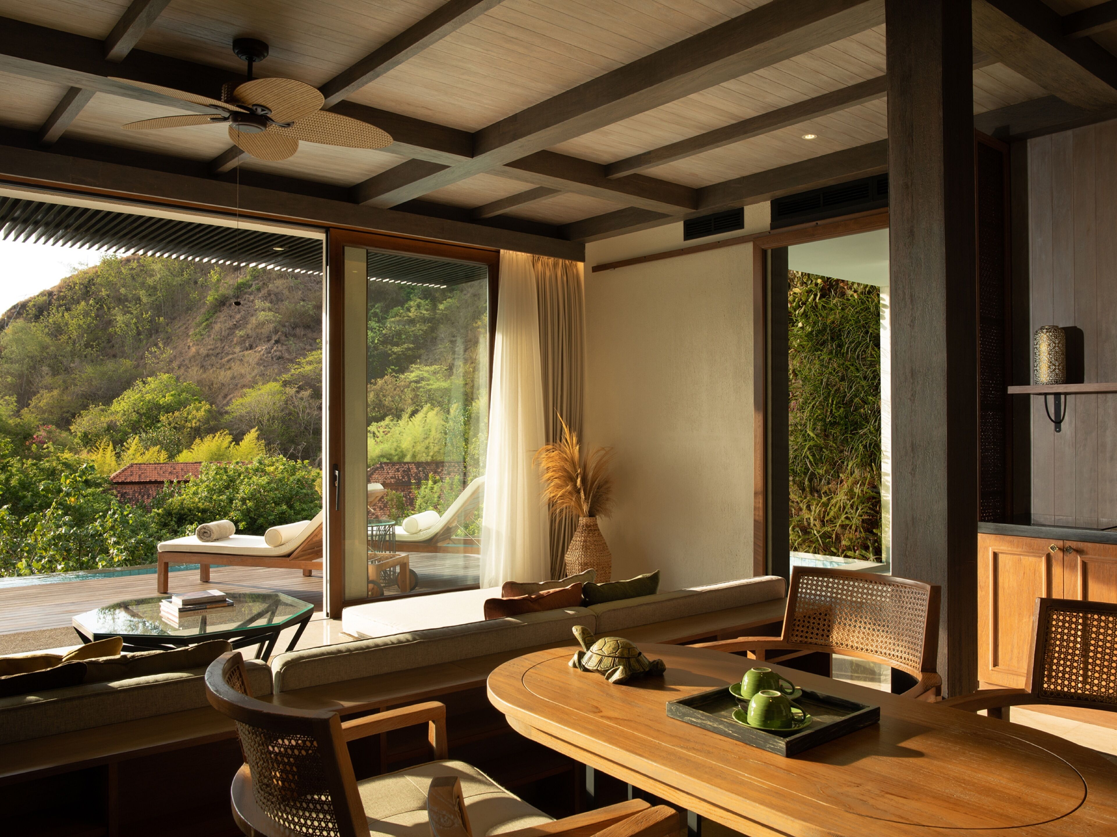 A modern dining area with a wooden table, chairs, and a view of a lush garden and hills.