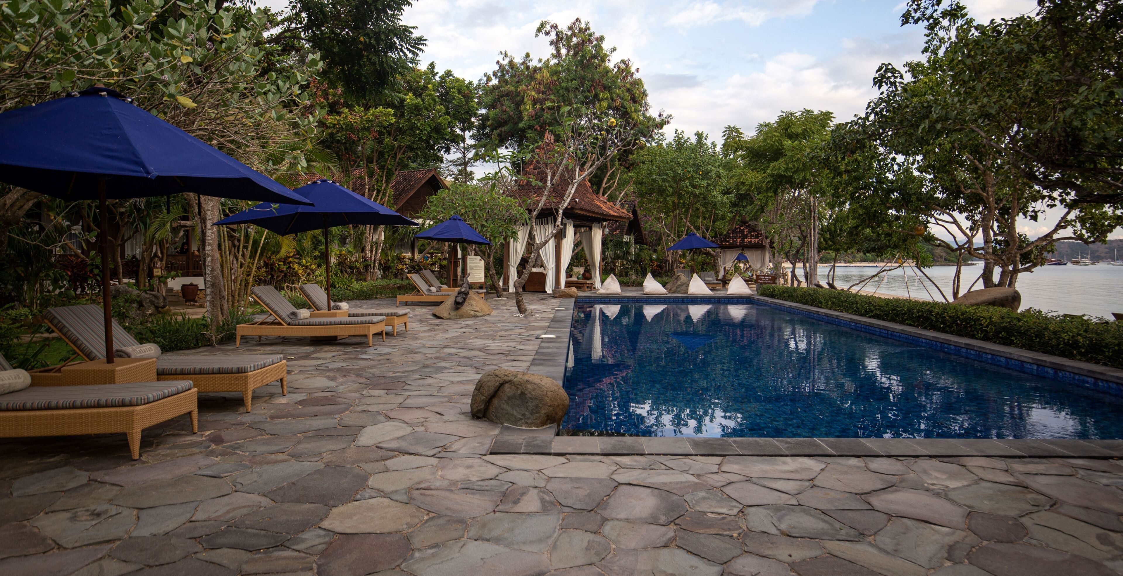 A pool area with lounge chairs and umbrellas, surrounded by trees and a view of the water.