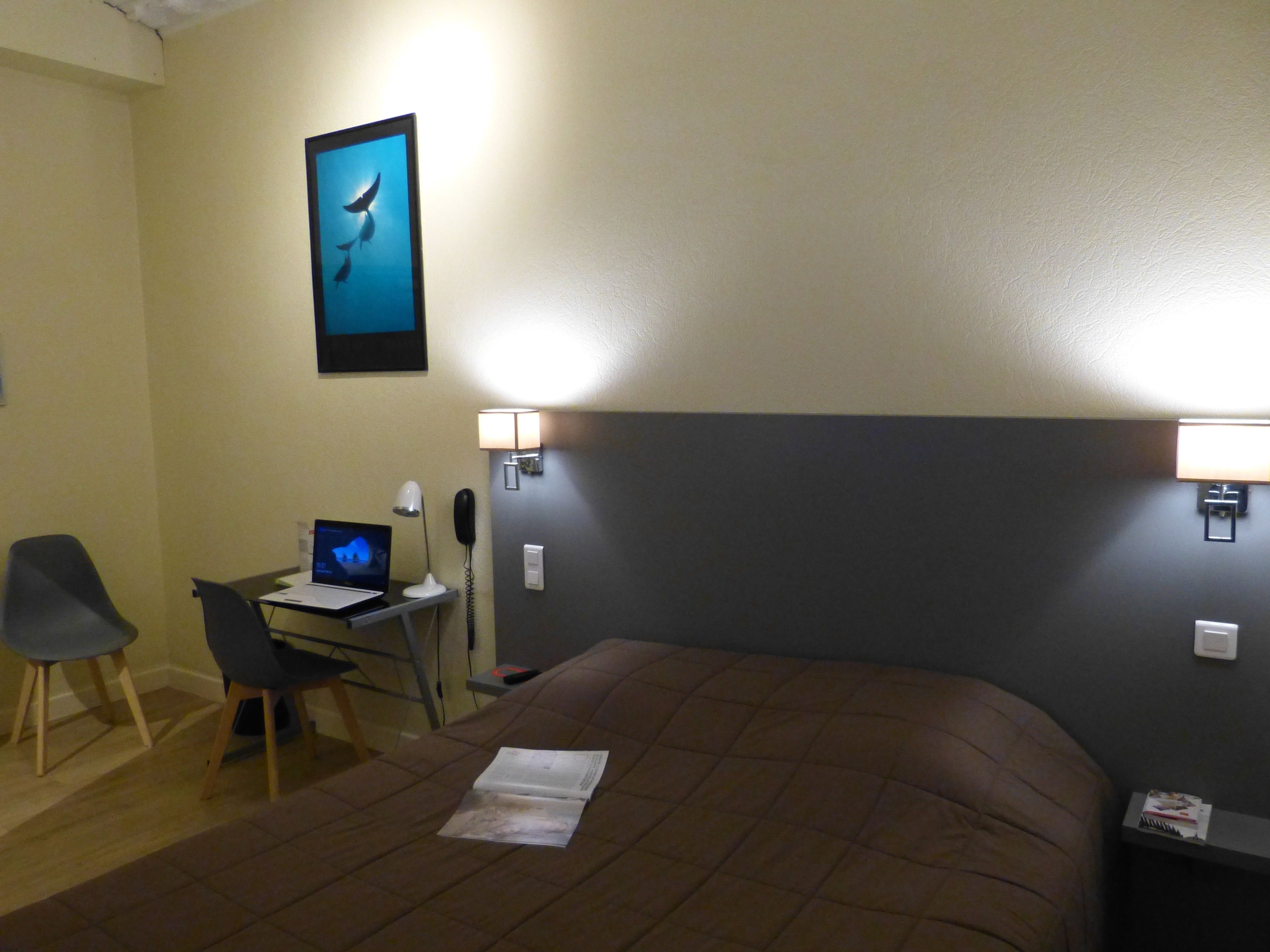 Une chambre d’hôtel comprenant un lit, un bureau avec un ordinateur portable, une chaise et deux lampes fixées au mur.