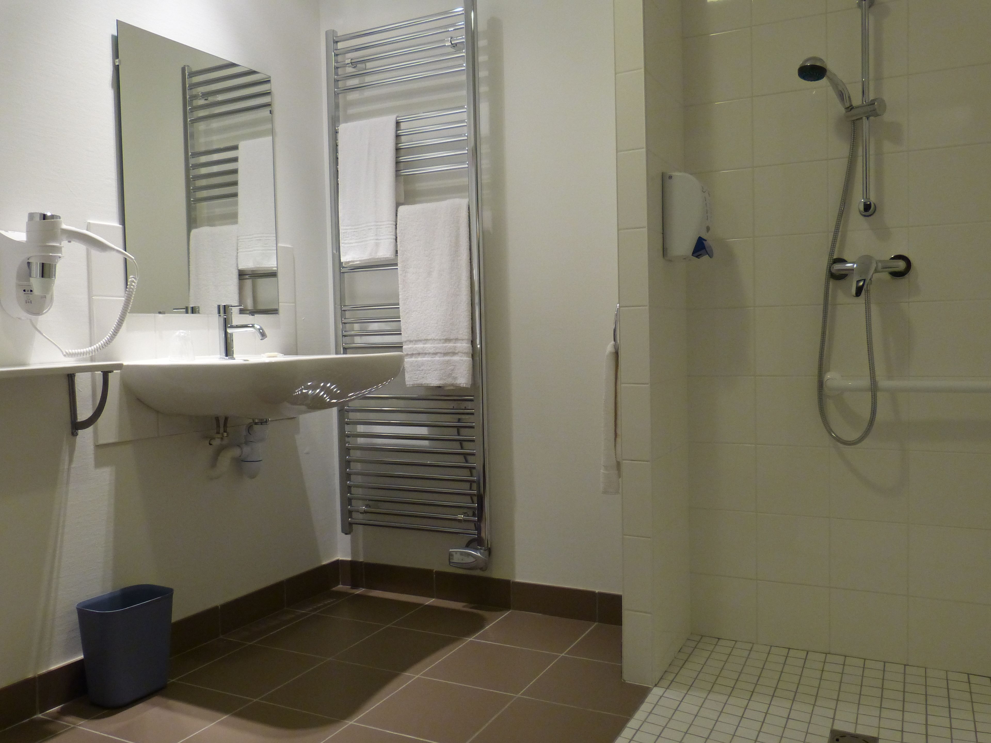 A bathroom with a sink, mirror, towel rack, shower, and a trash bin.