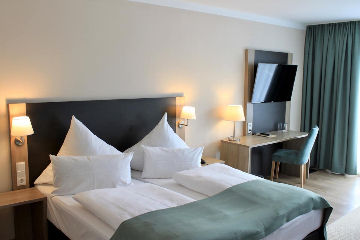 A hotel room with a large bed, two bedside tables with lamps, a desk with a chair, a flat-screen TV, and a window with curtains.