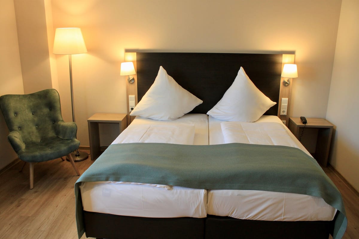 A hotel room with a large bed, two bedside lamps, a green armchair, and a bedside table.