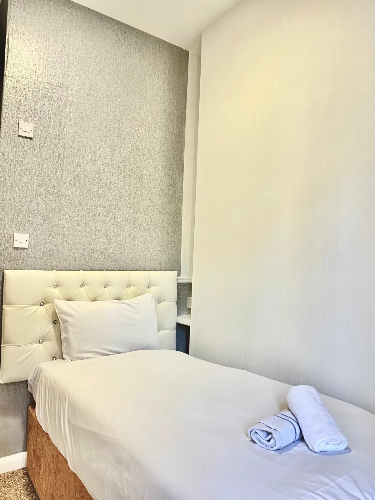 Een hotelkamer met een gestoffeerd hoofdeinde, een net opgemaakt bed met witte lakens en een opgerolde handdoek op het bed.