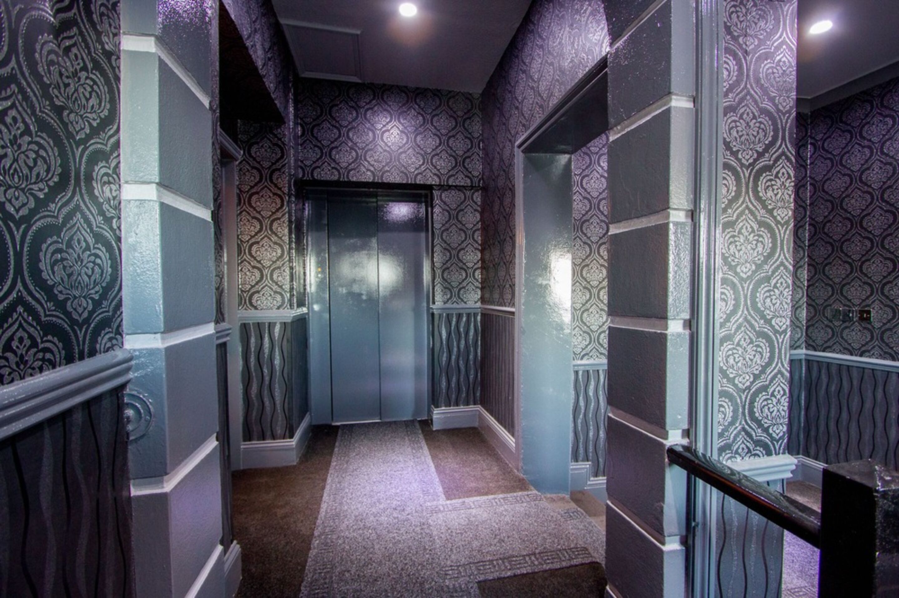 A hallway with patterned wallpaper, a carpeted floor, and a glass elevator.