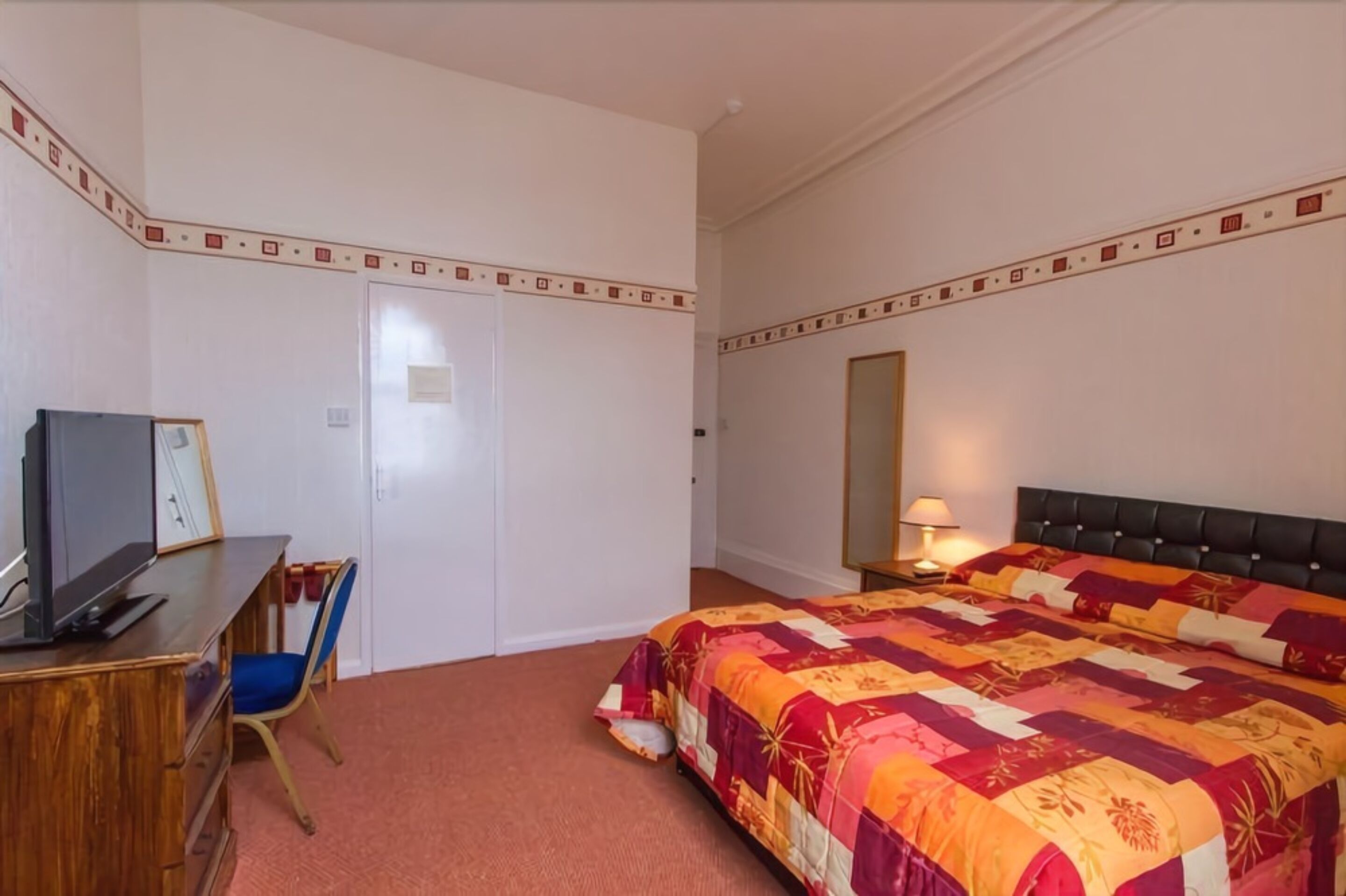 A hotel room with a bed, a desk with a computer, a chair, a wardrobe, and a mirror.