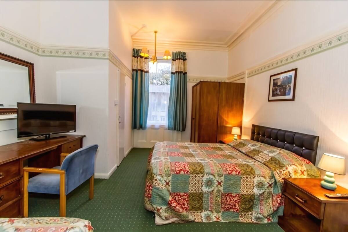 A hotel room with a bed, a desk with a television, a chair, a nightstand with a lamp, and a window with curtains.