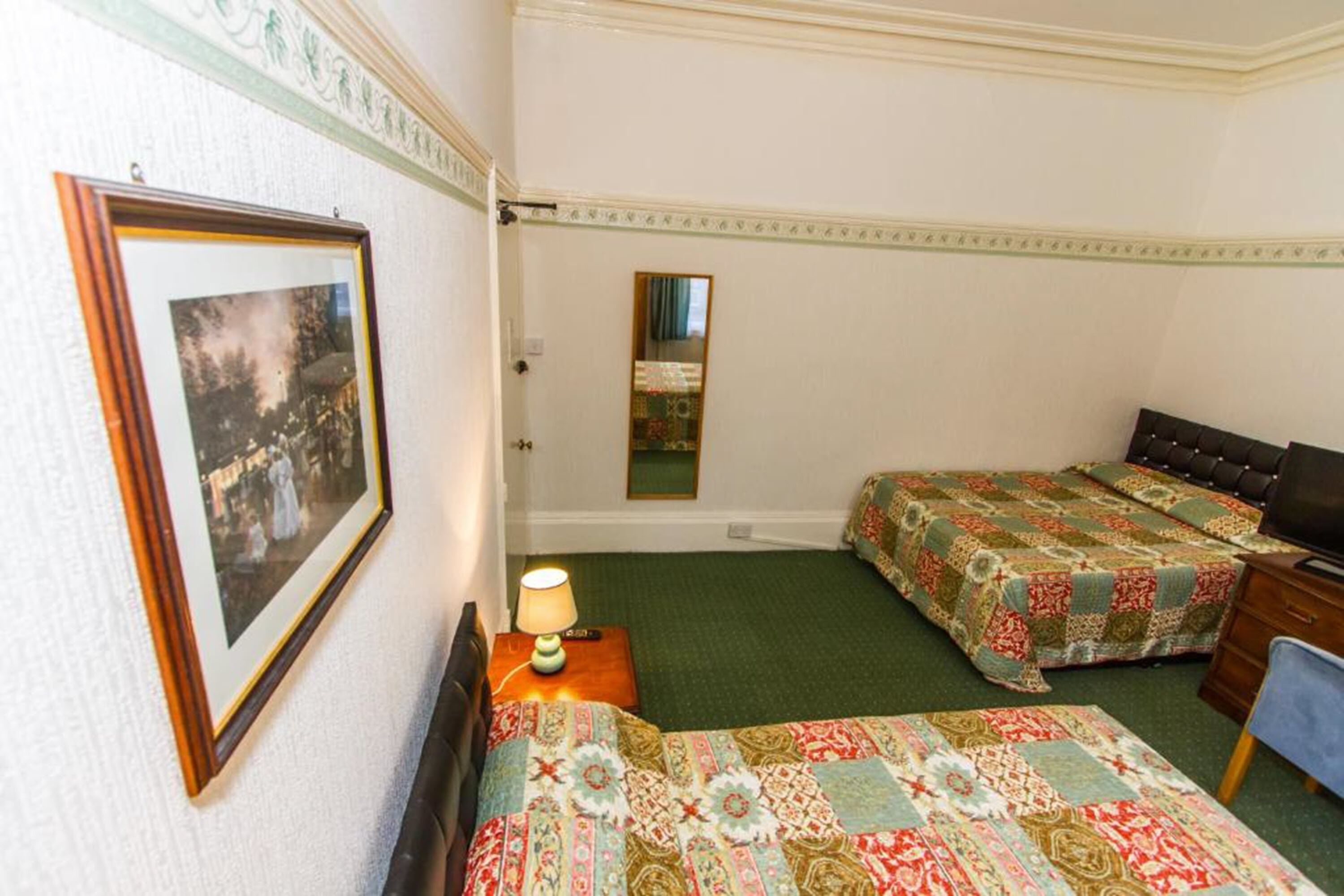 Habitación de hotel con dos camas individuales, una mesita de noche con lámpara, cómoda y un espejo. En la pared, se observa un cuadro enmarcado.