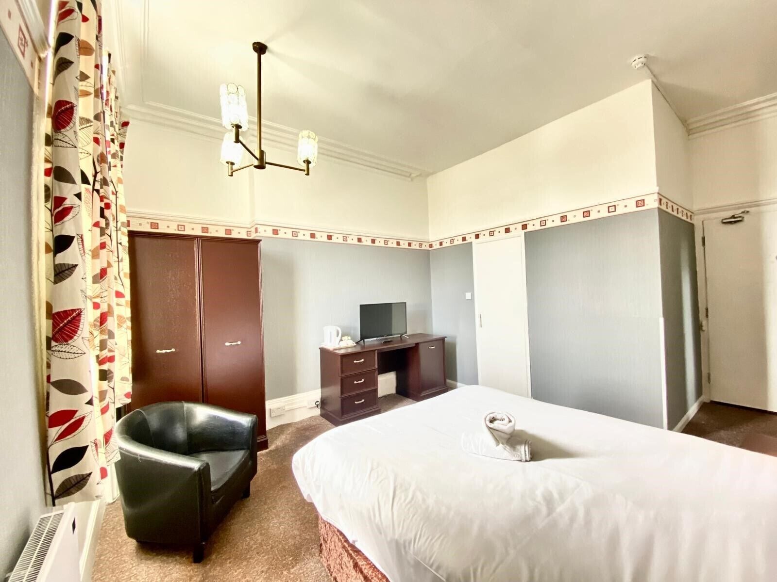 A hotel room with a bed, a desk with a TV, a wardrobe, a chair, and a ceiling light.