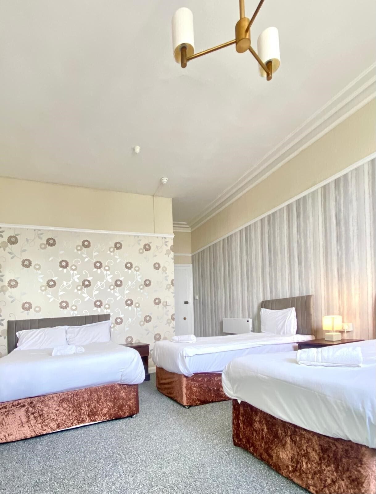 Una habitación de hotel con tres camas, un candelabro, papel tapiz con motivos florales y una cortina a rayas.