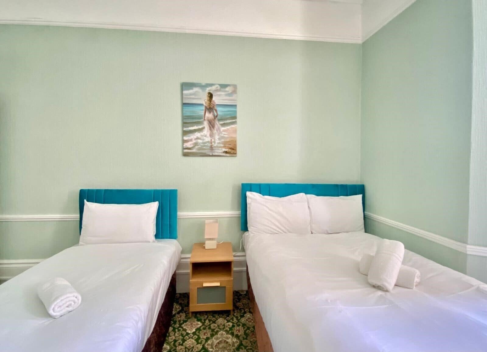 A hotel room with two beds, each with a blue headboard, white bedding, and a painting of a woman by the sea on the wall.