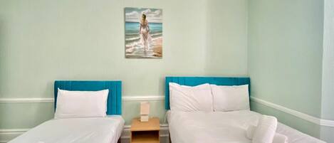 A hotel room with two beds, each with a blue headboard, white bedding, and a painting of a woman by the sea on the wall.