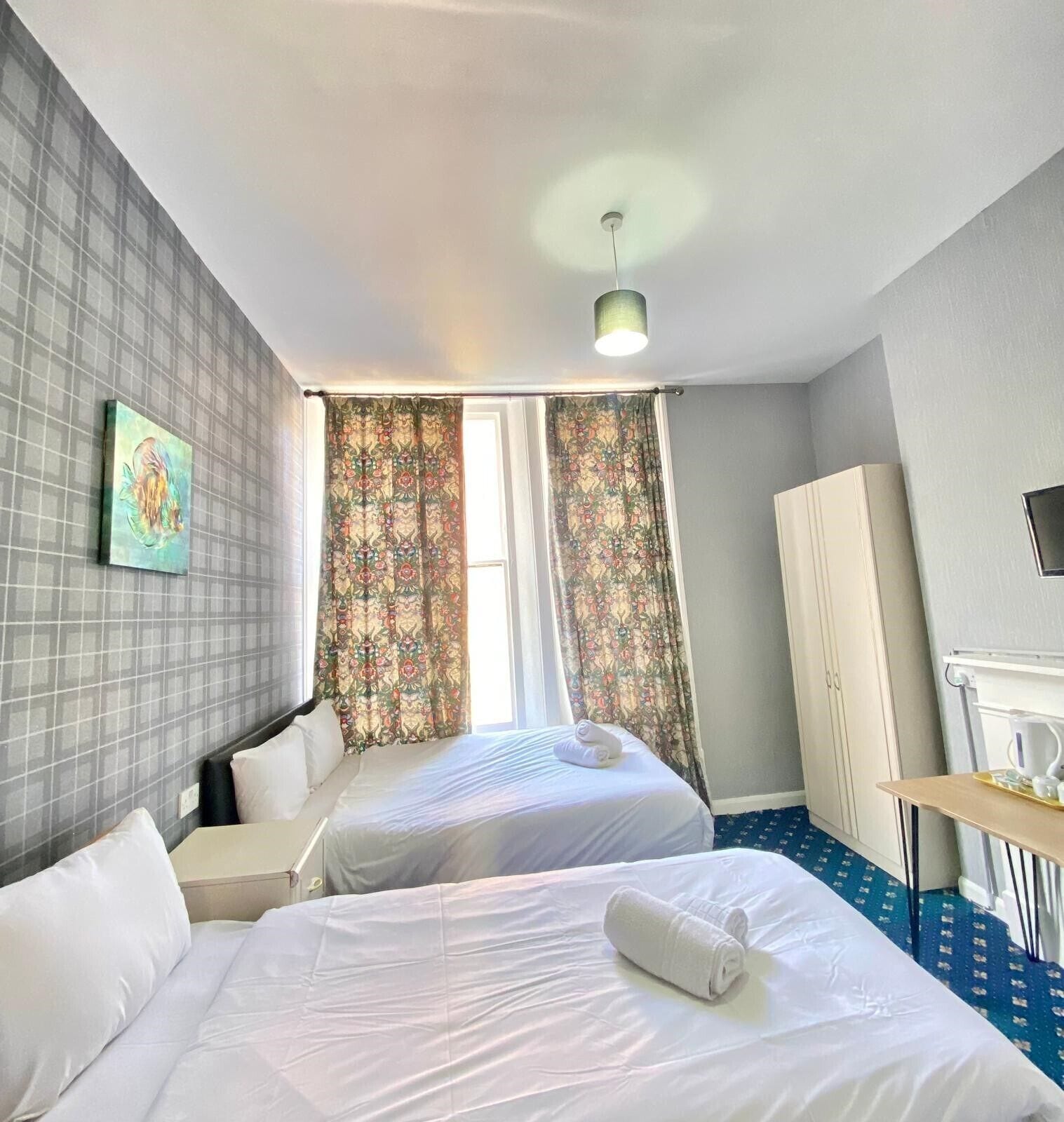 A hotel room with two beds, a large window with patterned curtains, a painting on the wall, and a desk with a cup and saucer.
