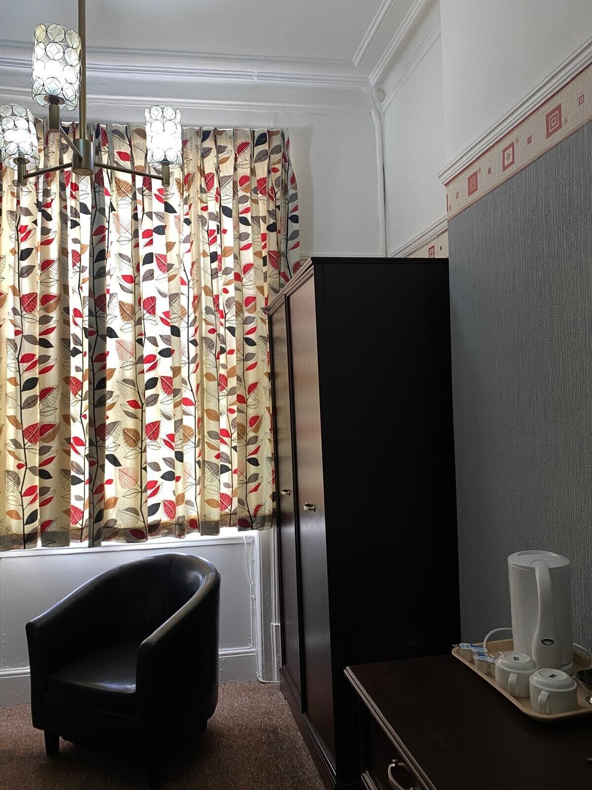Een hotelkamer met een groot raam, een zwarte fauteuil, een bureau met een koffiezetapparaat en een kast.