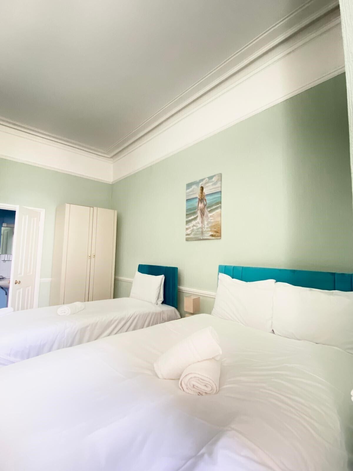 A hotel room with two beds, a painting of a woman on the beach, and a view of the sea through an open door.