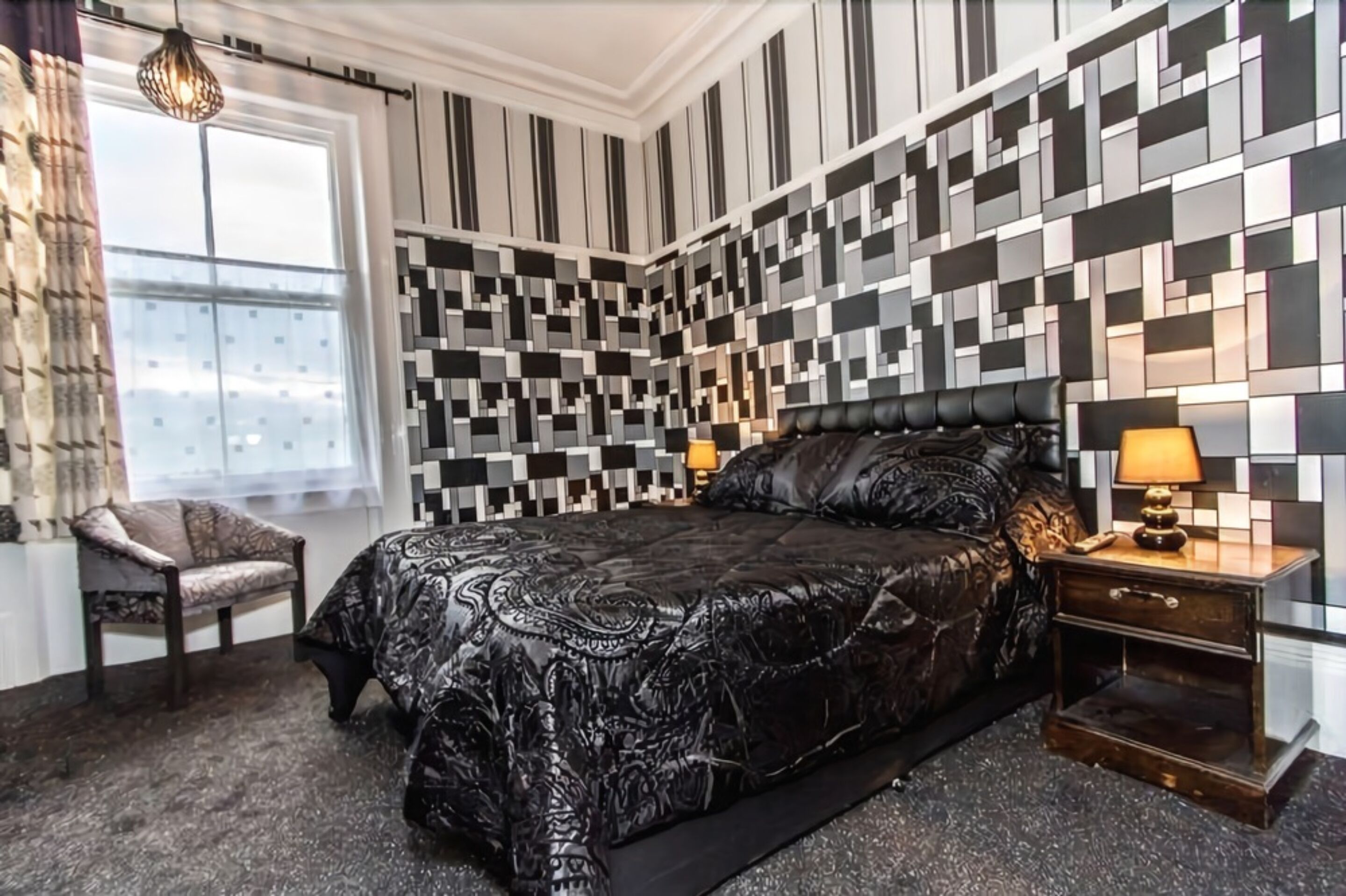 Une chambre moderne avec une couvre-lit à motifs, une tête de lit en cuir noir, une table de chevet avec une lampe et un fauteuil.
