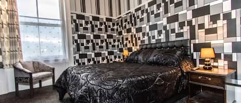Une chambre moderne avec une couvre-lit à motifs, une tête de lit en cuir noir, une table de chevet avec une lampe et un fauteuil.