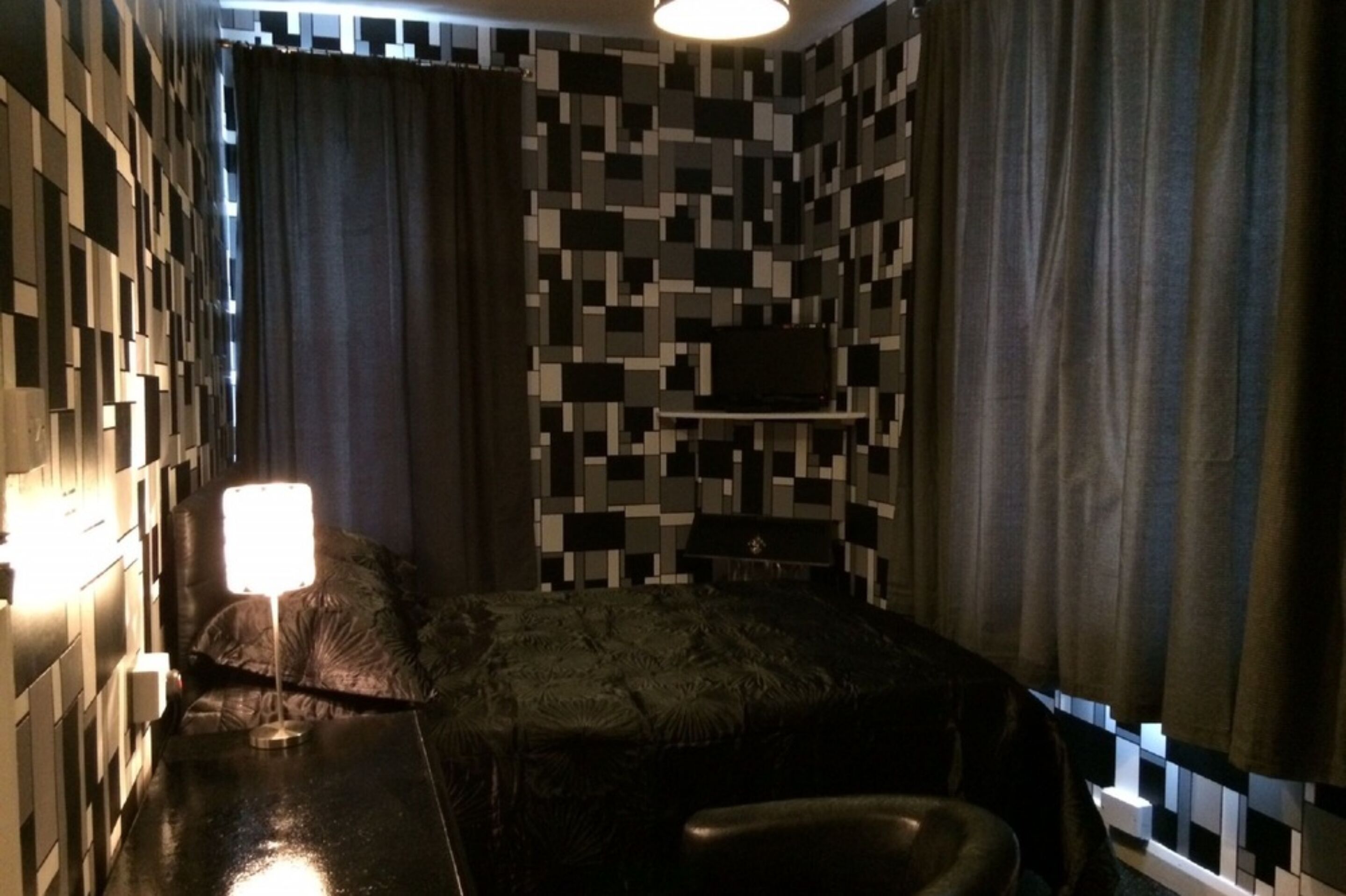 Une pièce comprenant un lit, une petite table, une lampe et un mur orné d’un motif pixélisé.