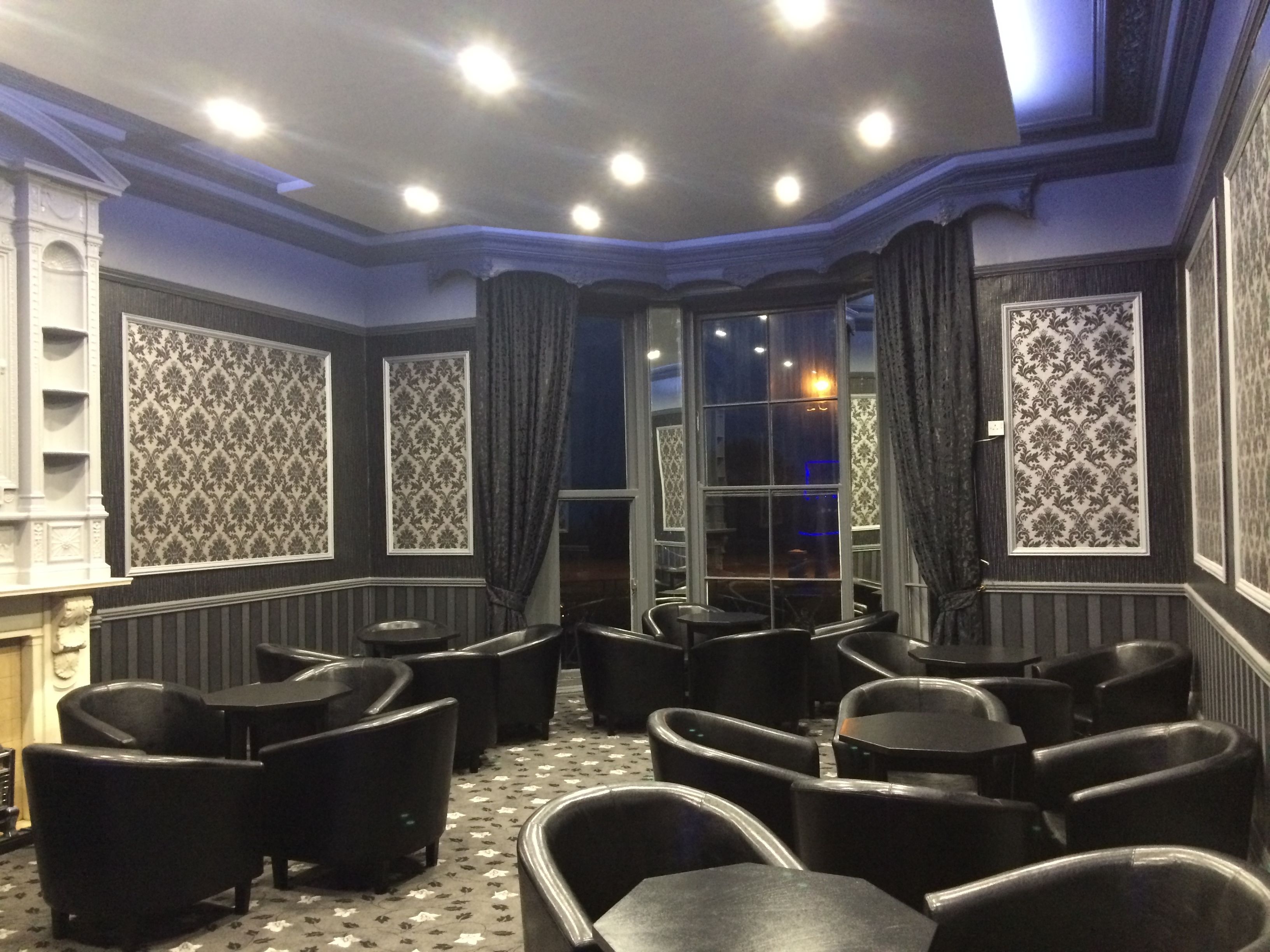 A corner room with a fireplace, patterned wallpaper, and a seating area with tables and chairs.