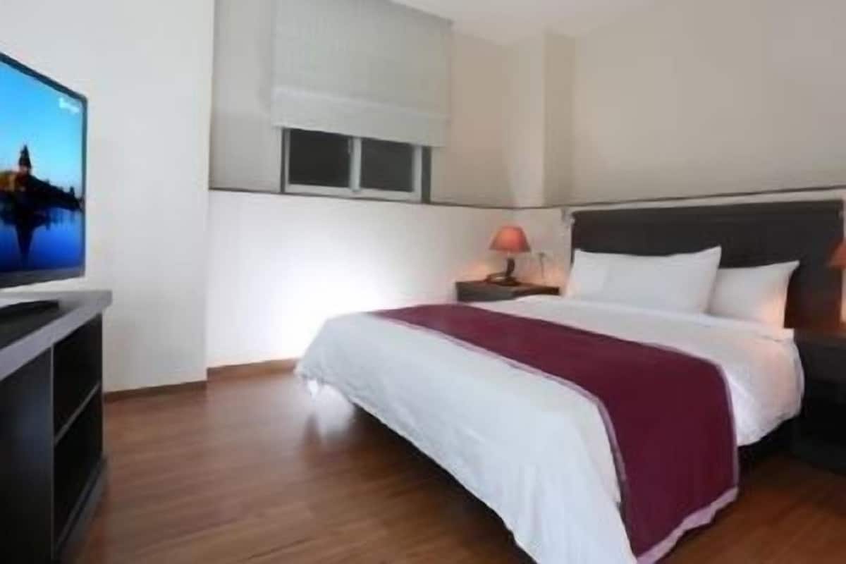 A hotel room with a bed, a TV on a stand, a window, and a nightstand with a lamp.