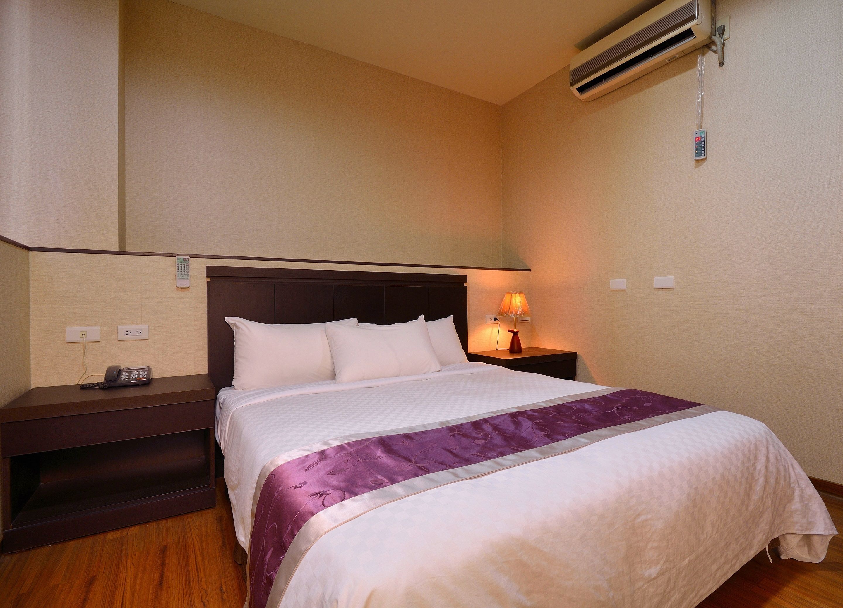 Una cama bien hecha con una colcha blanca y morada, una mesita de noche de madera con una lámpara y un aire acondicionado instalado en la pared.