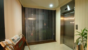 Un moderno ascensor con detalles metálicos, un tabique de vidrio con persianas verticales y un distribuidor de periódicos con revistas.
