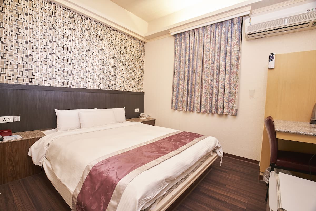 A hotel room with a neatly made bed, a wooden headboard, a desk with a chair, and an air conditioning unit on the wall.