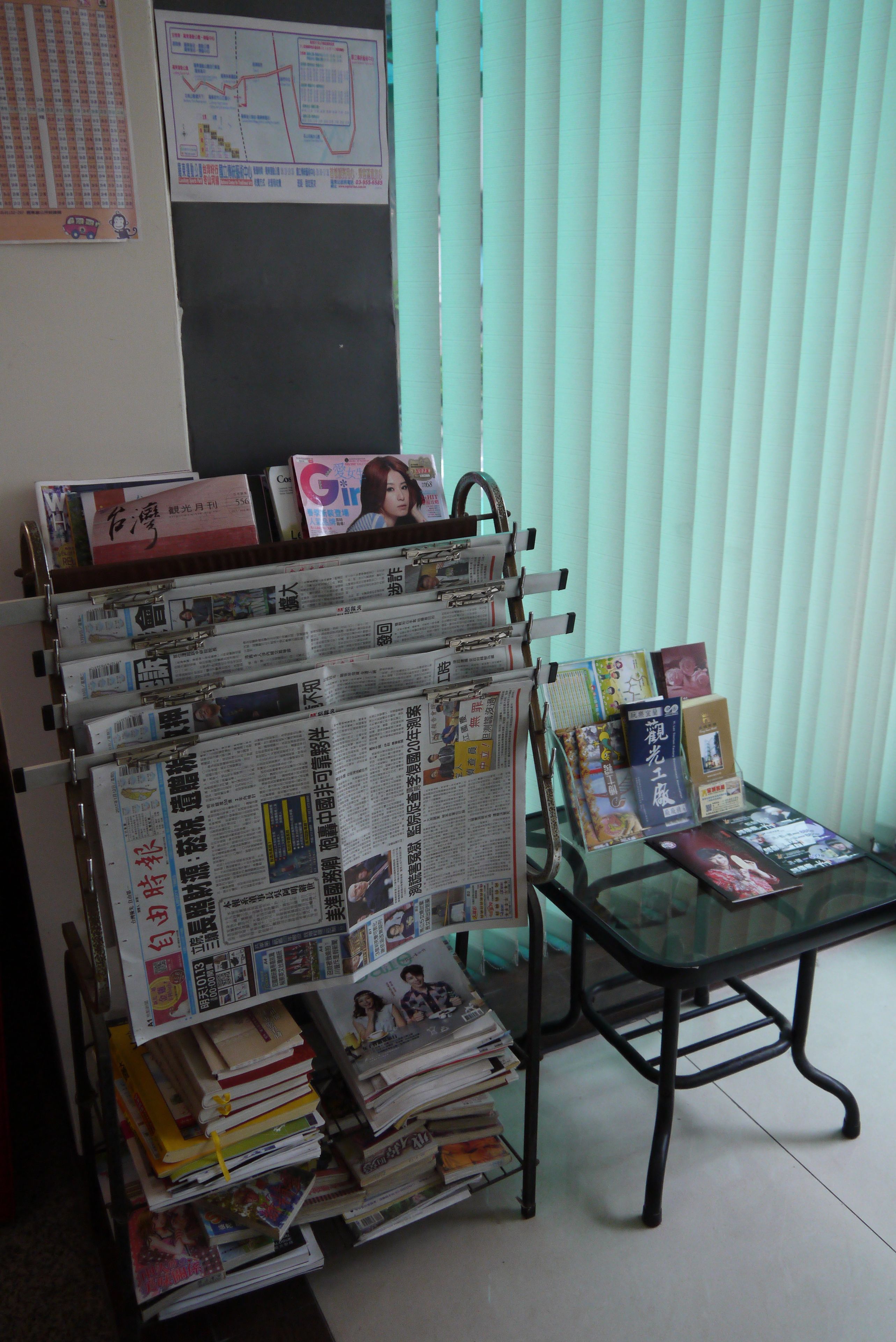 Una habitación con una mesa de vidrio que muestra revistas y libros, un tablón de anuncios con papeles y un mapa, y una ventana con persianas.