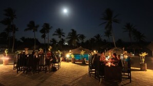 An outdoor dining area with tables and chairs, a pool, and palm trees under a night sky with a visible moon. - Blue Moon Beach Holiday Resort (Jangamo)