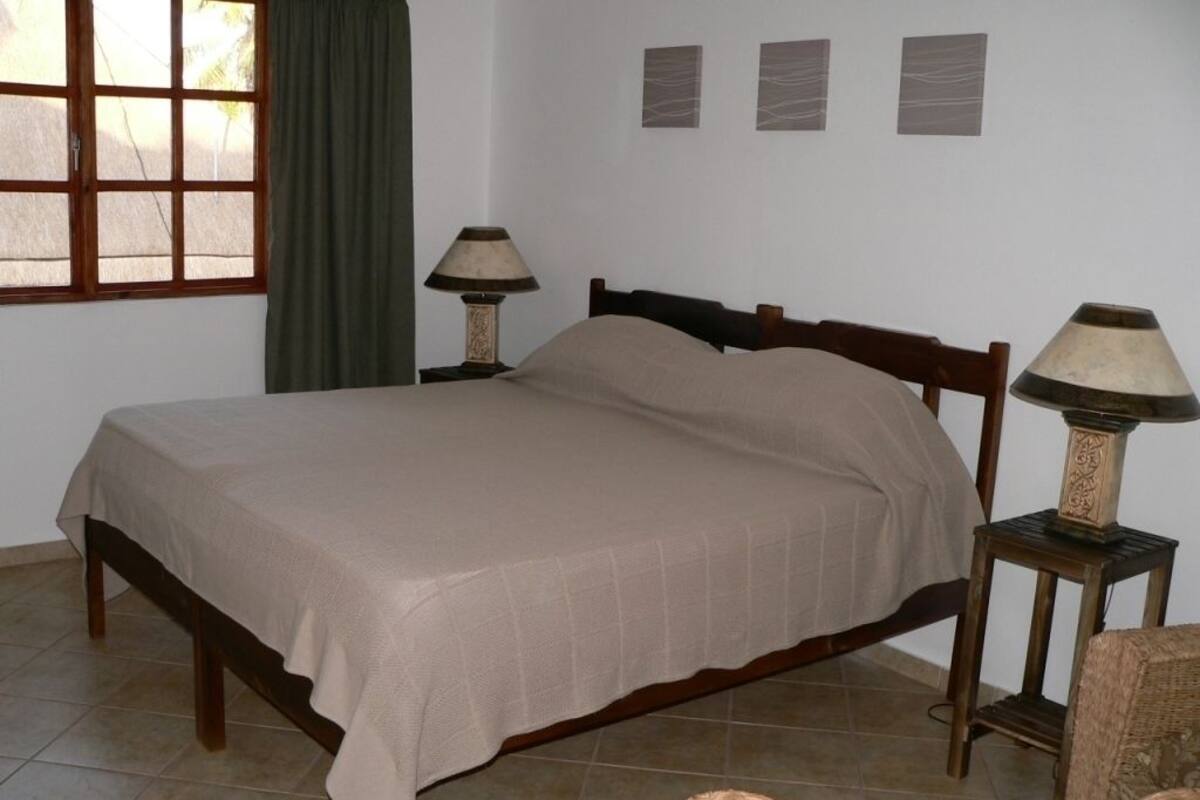 A bedroom with a bed, two bedside tables with lamps, a window with curtains, and three wall-mounted picture frames.