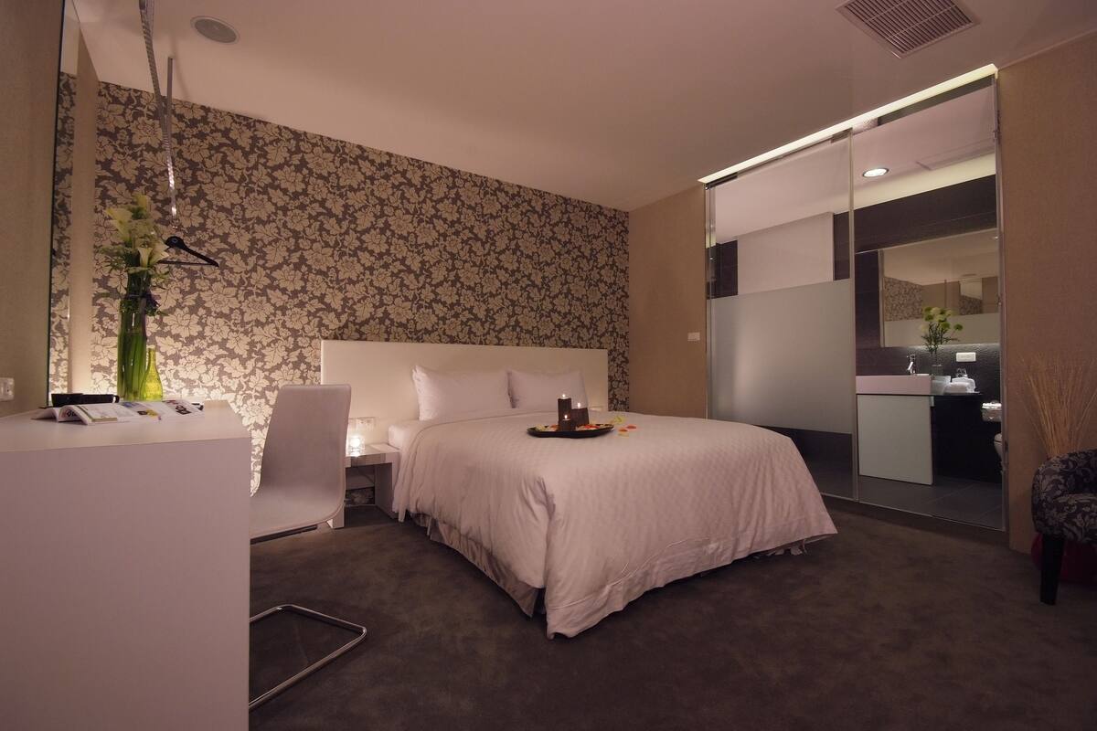 A modern hotel room with a large bed, a desk, and a small kitchenette.