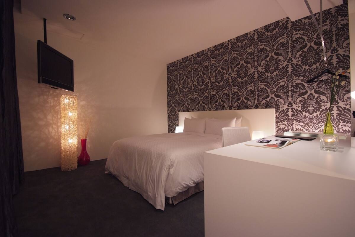 A modern hotel room with a large bed, a flat-screen TV mounted on the wall, a decorative light fixture, and a patterned wallpaper on the upper wall.
