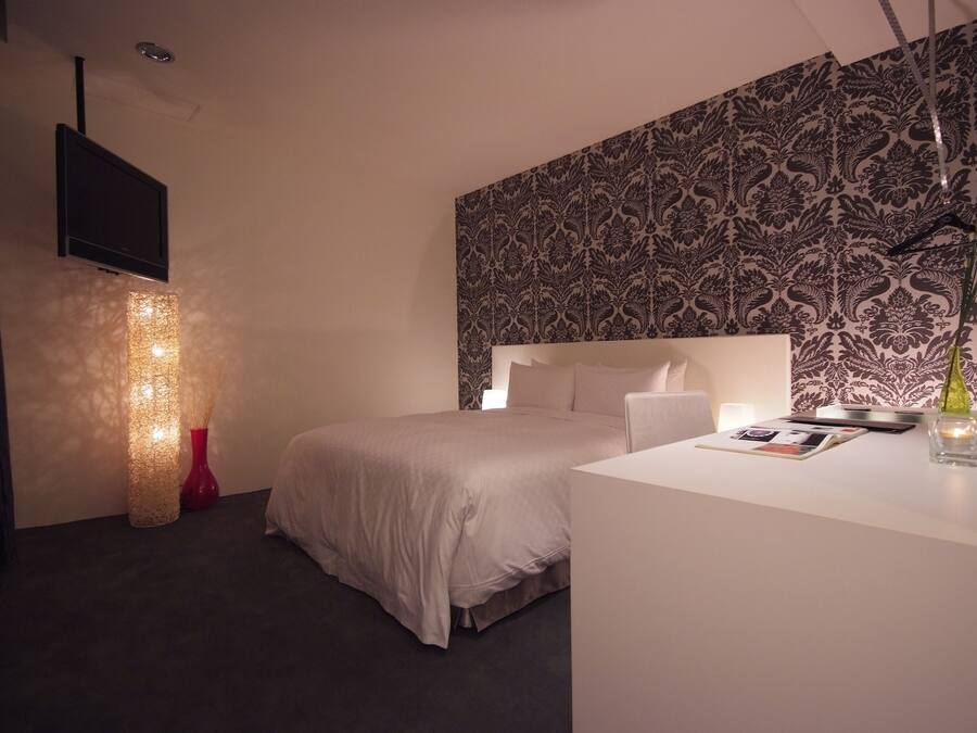 Een moderne hotelkamer met een groot bed, een flatscreen televisie aan de muur, een decoratieve lamp en behang met een patroon op het bovenste deel van de muur.