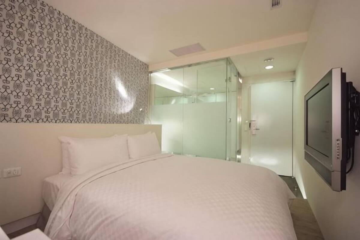 A hotel room with a bed, a television, and a glass shower enclosure.