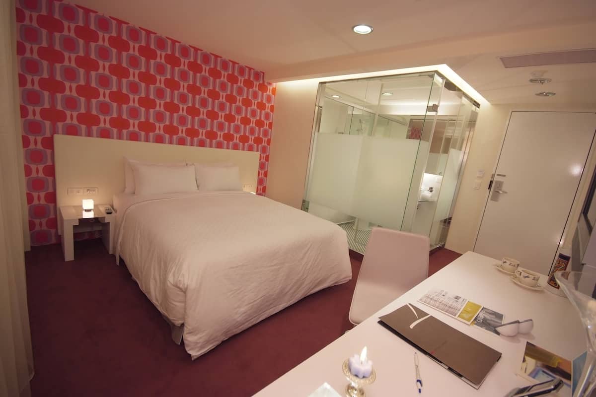 A hotel room with a large bed, a desk with a laptop, a chair, a nightstand, and a glass-enclosed shower area.