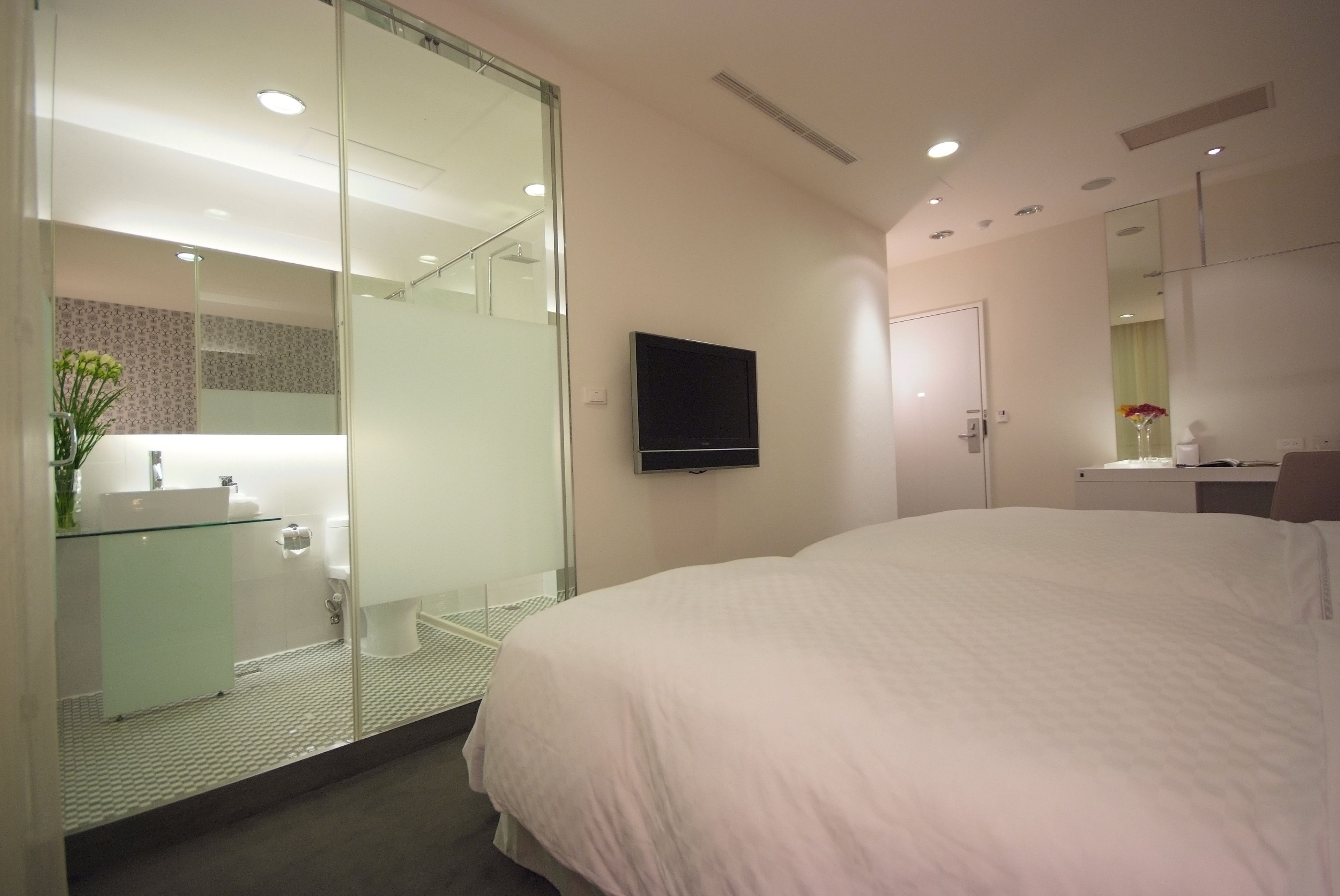 Habitación de hotel moderna con una cama grande, cabina de ducha de vidrio, televisor montado y un pequeño escritorio con espejo.