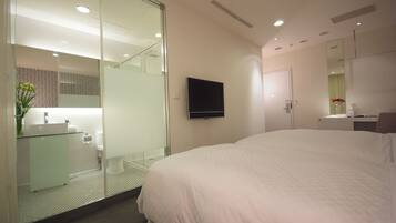 Habitación de hotel moderna con una cama grande, cabina de ducha de vidrio, televisor montado y un pequeño escritorio con espejo.