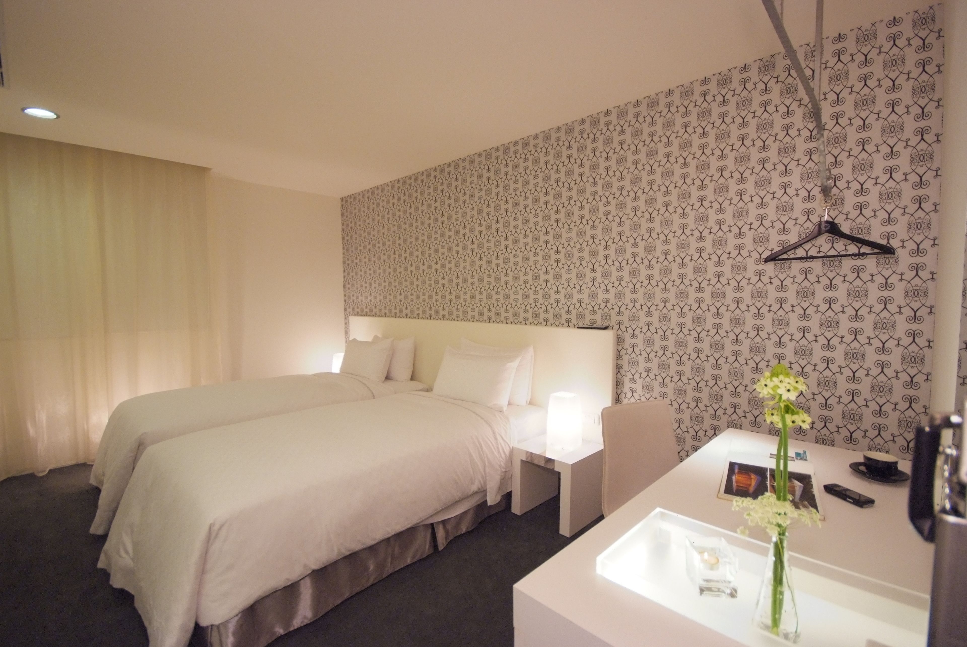 A hotel room with two beds, a white bedside table, a mirror, a hanging black hanger, and a vase with flowers.