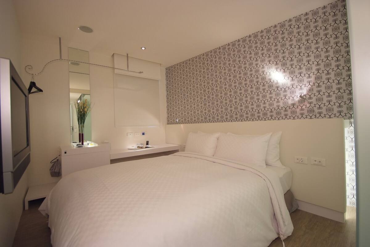 A modern bedroom with a large bed, a bedside table, a mirror, and a patterned wall.
