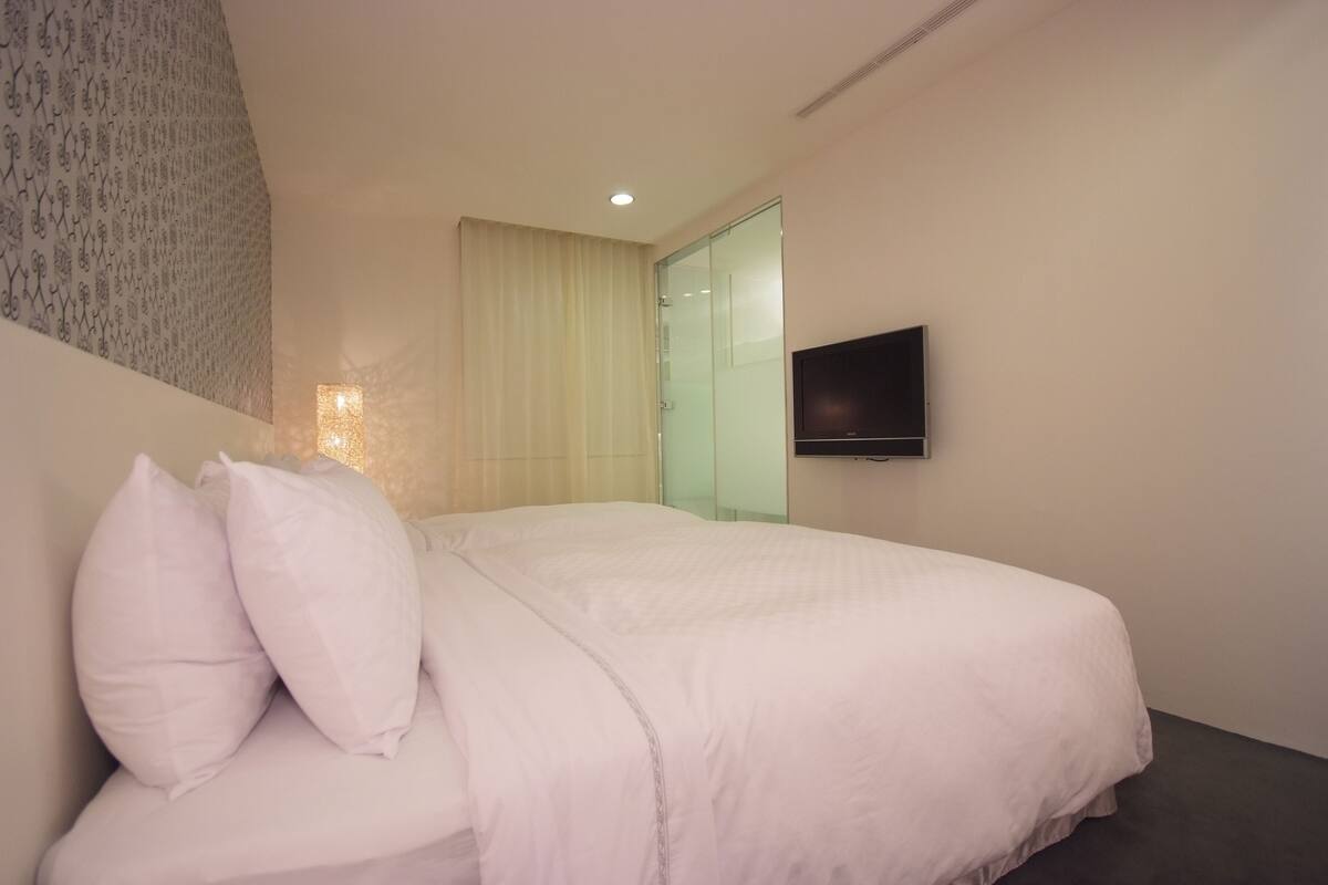 A modern hotel room with a large bed, a flat-screen TV mounted on the wall, and a glass door leading to a bathroom.