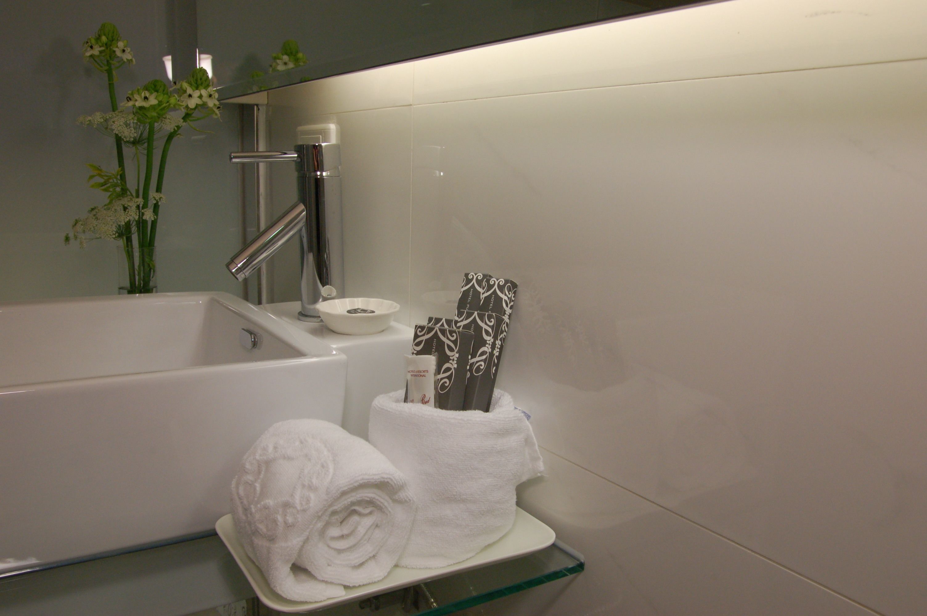 Un baño con bañera, repisa de vidrio con toallas, espejo y un florero con flores.
