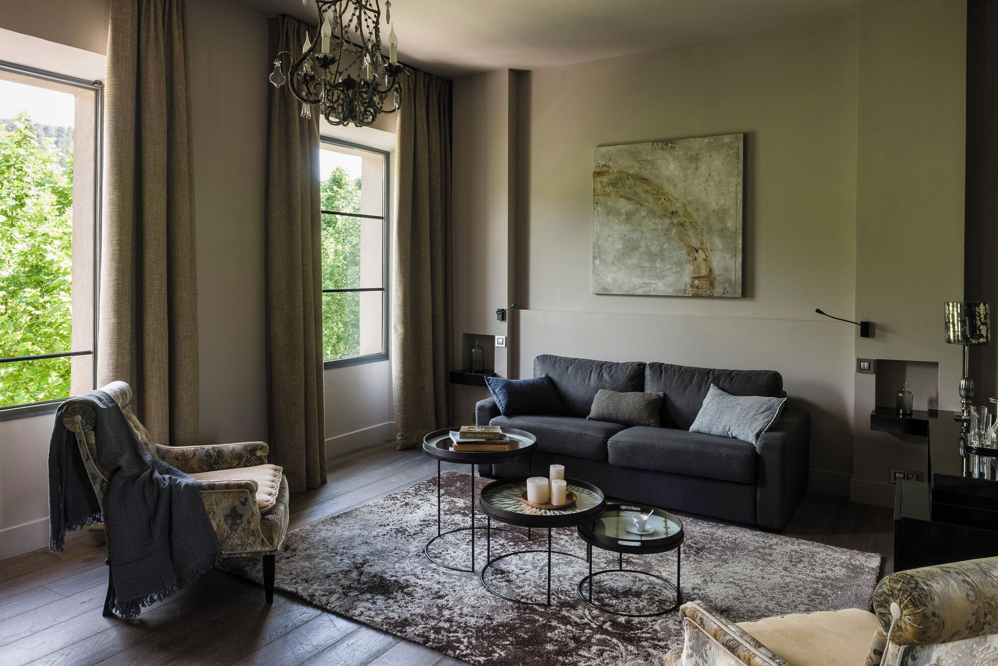 A living room with a sofa, armchair, and a chandelier. There is a window with curtains, a painting on the wall, and a lamp.