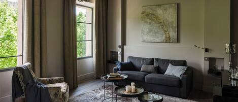A living room with a sofa, armchair, and a chandelier. There is a window with curtains, a painting on the wall, and a lamp.