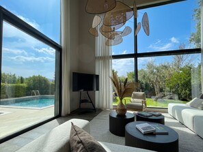 A modern living room with a large window, a flat-screen TV, a sofa, a coffee table, and a view of a swimming pool. - Les Lodges Sainte-Victoire Hotel & Spa (Le Tholonet)