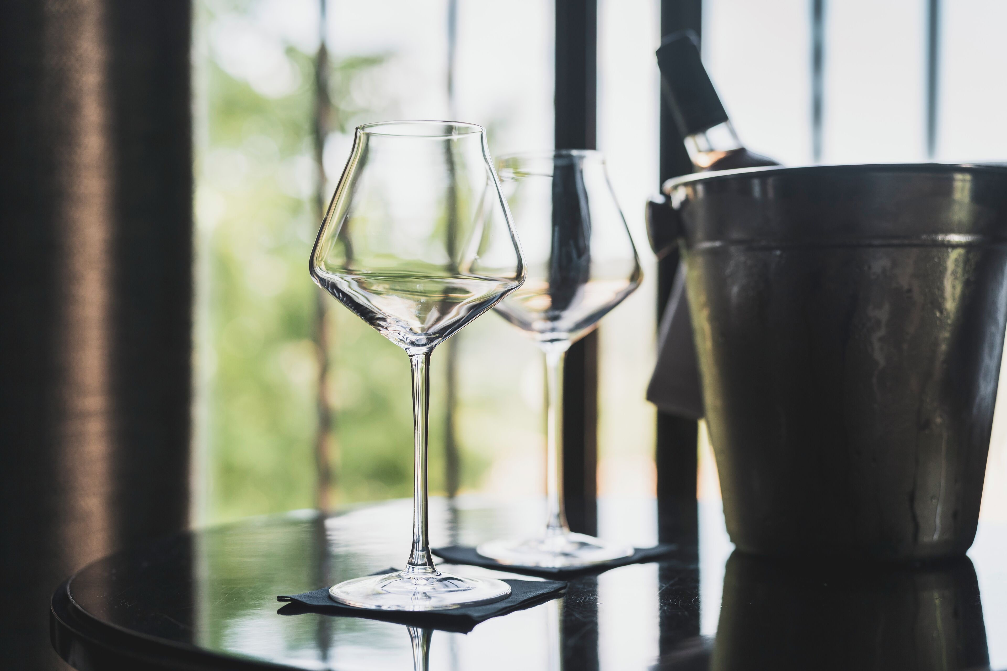 Una mesa con dos copas de vino y un balde metálico, probablemente para hielo, en una habitación bien iluminada con vistas al exterior.