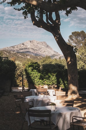 An outdoor dining area with tables and chairs, a large tree, and a mountain in the background. - Les Lodges Sainte-Victoire Hotel & Spa (Le Tholonet)