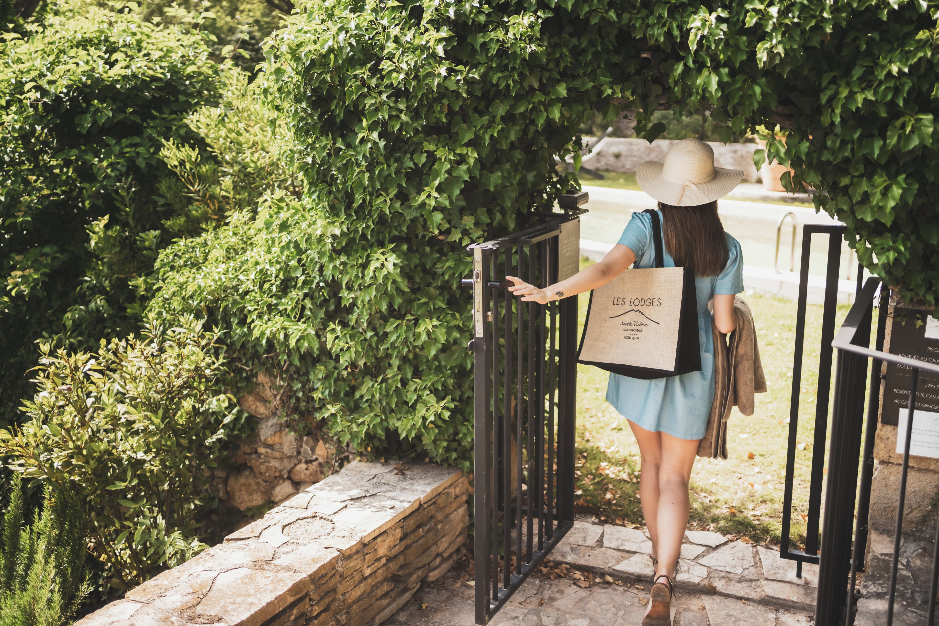 A person exiting a gate with a shopping bag, surrounded by greenery and stone walls.