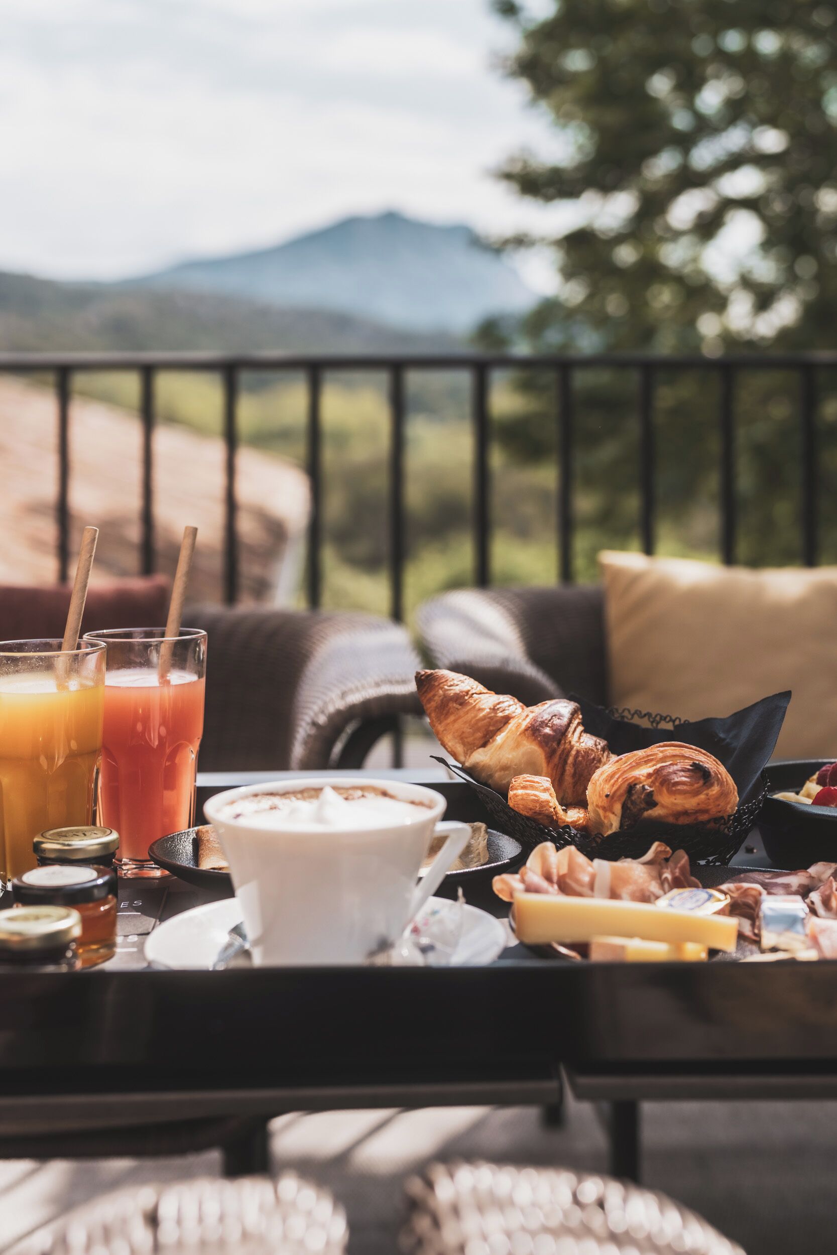 Une table de petit-déjeuner avec des croissants, une tasse de café et des verres de jus, le tout sur un balcon donnant sur un paysage montagneux.