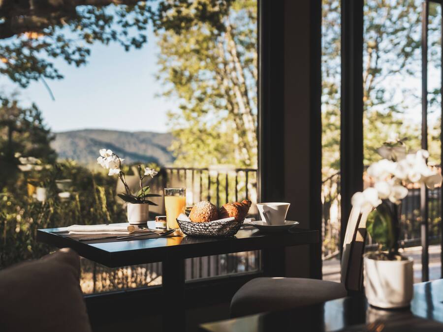 A table with a bowl of croissants, a cup, and a glass of orange juice, with a view of trees and mountains through large windows.